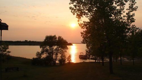 Sunset over a calm lake, with trees in silhouette and a boat docked along the shore.