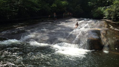 A rocky riverbed with a small waterfall, surrounded by lush greenery and people enjoying the water.