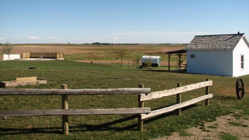 A serene rural landscape featuring a small white house, a wooden fence, and expansive fields under a clear blue sky.