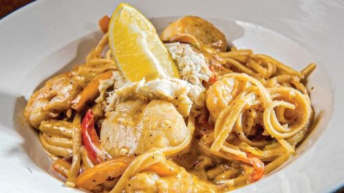 A plate of pasta with seafood, topped with crab meat and a lemon wedge, garnished with colorful bell peppers.