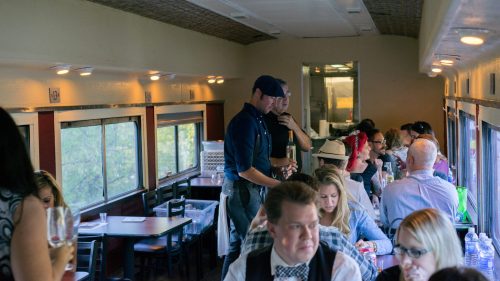 A dining car interior with patrons at tables, a waiter serving drinks, and large windows showing a scenic view outside.