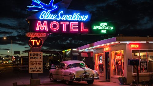 Neon sign of Blue Swallow Motel at dusk, featuring a classic car parked outside and a retro vibe.