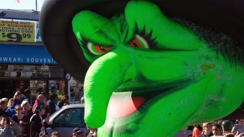 A large green witch balloon with a prominent nose and hat, looming over a crowd at a festive event.