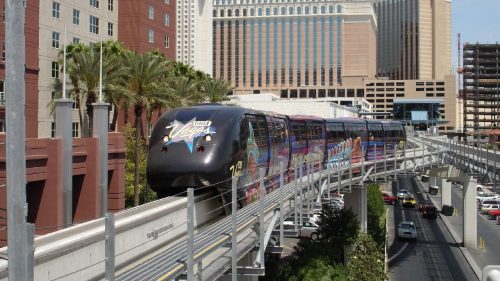 A monorail travels above a busy street, with hotels and palm trees in the background under a clear blue sky.