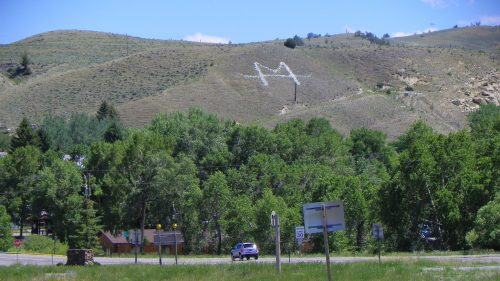 A large white letter "M" is painted on a hillside, surrounded by green trees and a clear blue sky.