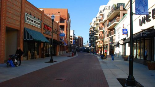 A wide, sunny street lined with shops and apartments, featuring pedestrians and outdoor seating.