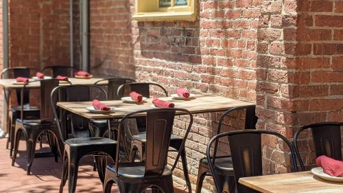 Outdoor dining area with empty tables and black chairs against a brick wall, set with red napkins.
