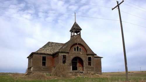 Abandoned wooden building with a steeple, overgrown grass, and a power line against a cloudy sky.