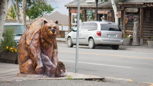 A life-sized bear sculpture sits on a sidewalk near a street, with shops and trees in the background.
