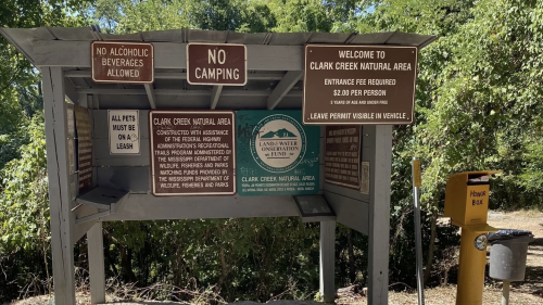 Signage at Clark Creek Natural Area with rules, entrance fee info, and pet policy. Surrounded by greenery.