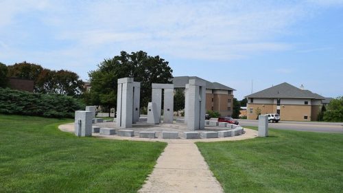 A stone monument with tall pillars surrounded by grass and a walkway, near residential buildings under a blue sky.