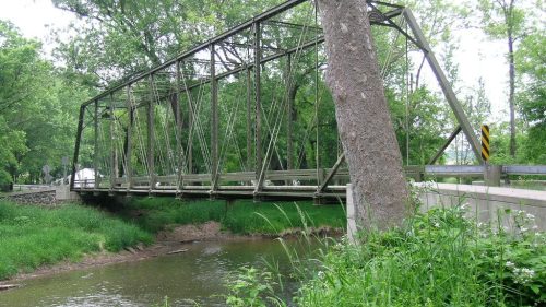 A metal truss bridge spans a small creek, surrounded by lush greenery and trees.