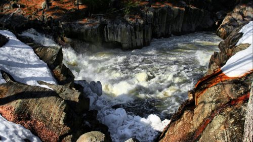Rushing river water flows through rocky terrain, surrounded by snow and trees in a natural landscape.