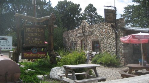 A rustic stone building with a sign for Pine Creek General Store and Restaurant, surrounded by trees and picnic tables.