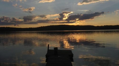 A serene lake at sunset, with clouds reflecting on the water and a wooden dock extending into the scene.