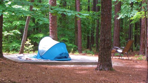A blue tent set up on a concrete pad in a forest, surrounded by tall trees and a camping chair nearby.