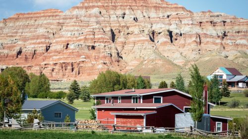 A red-roofed house in a green field with a colorful, layered mountain in the background under a blue sky.