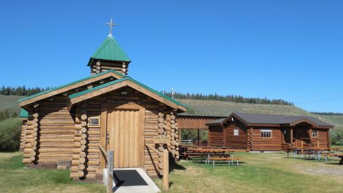 A rustic log cabin church with a green roof, surrounded by trees and a grassy area, alongside another wooden building.