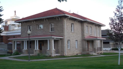 Historic stone building with a red tile roof, surrounded by green grass and trees, in a serene outdoor setting.