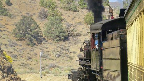 A vintage steam train chugs along a scenic route, releasing black smoke against a backdrop of hills and trees.