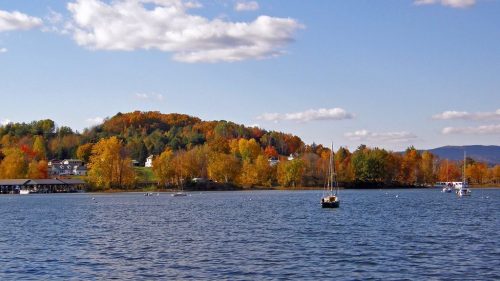 A serene lake scene with colorful autumn trees, a small sailboat, and fluffy clouds in a clear blue sky.