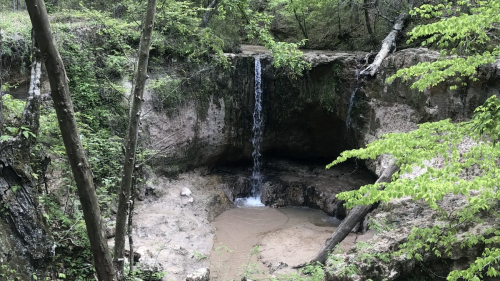 A serene waterfall cascades into a sandy pool, surrounded by lush green trees and rocky terrain.