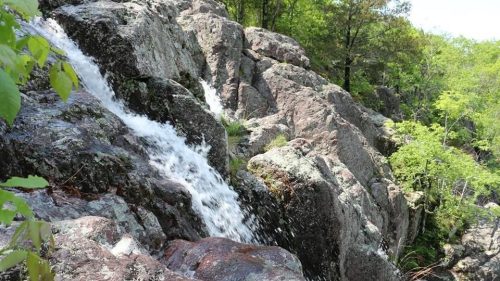 A small waterfall cascades over rocky terrain, surrounded by lush green trees and foliage.
