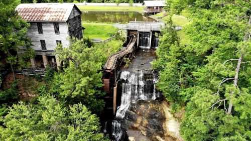 Aerial view of a rustic mill beside a waterfall, surrounded by lush greenery and a calm pond in the background.