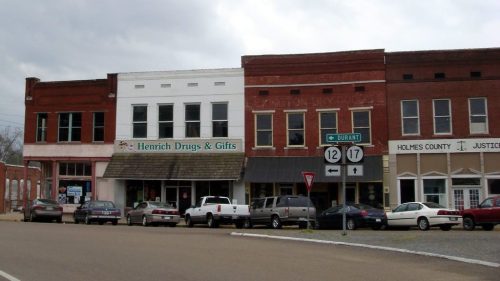 A small town street featuring brick storefronts, including Henrich Drugs & Gifts, with parked cars and cloudy skies.