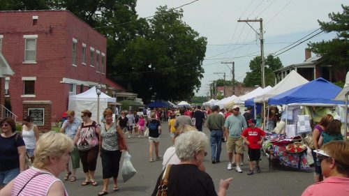 A bustling street market with people browsing tents and stalls, surrounded by trees and buildings on a sunny day.