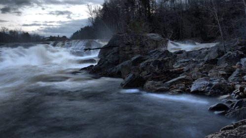 A turbulent river flows over rocks under a cloudy sky, surrounded by trees and mist.
