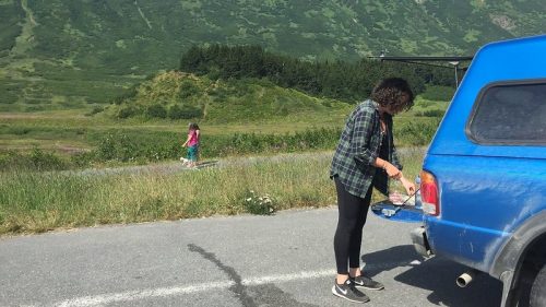 A person prepares food by a blue truck in a grassy area, with a mountain backdrop and another person walking in the distance.