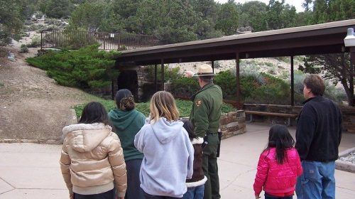 A group of people, including a park ranger, stands near a visitor center surrounded by trees and a hillside.