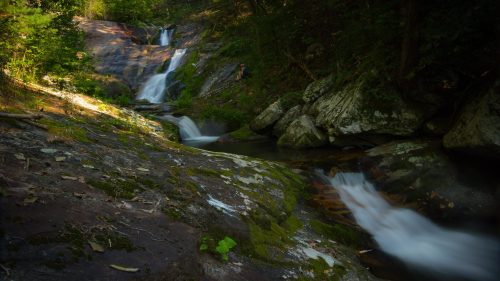 A serene waterfall cascades over rocky terrain, surrounded by lush greenery and dappled sunlight.
