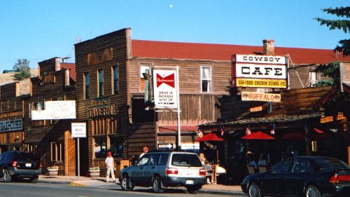 A street view of rustic buildings, including Cowboy Café and a Budweiser sign, under a clear blue sky.
