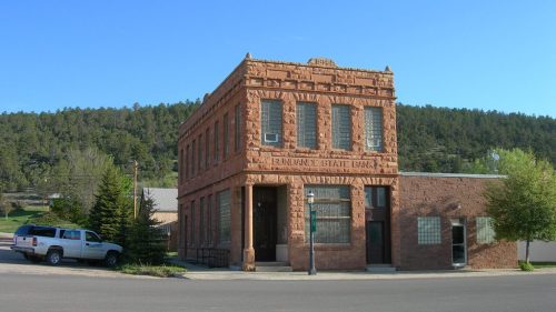 Historic brick building with ornate details, surrounded by trees and parked vehicles in a small town setting.