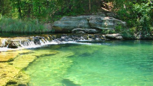 A serene, clear blue-green stream flows over rocks, surrounded by lush greenery and trees.