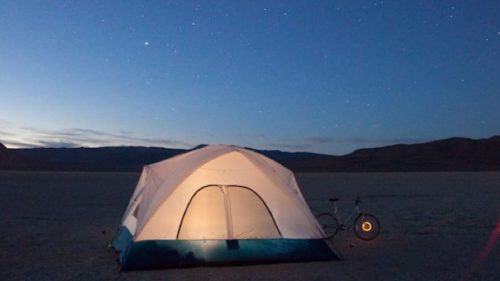 A tent illuminated at night under a starry sky, with a bicycle nearby on a barren landscape.