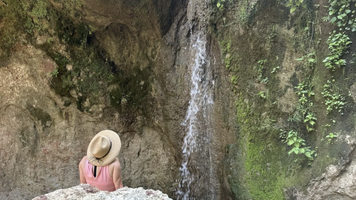 A person in a hat sits on a rock, gazing at a small waterfall surrounded by mossy cliffs.