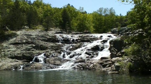 A serene waterfall cascades over rocky terrain, surrounded by lush green trees under a clear blue sky.
