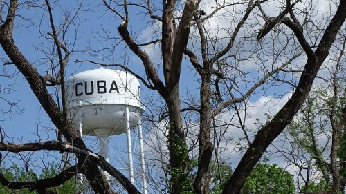 A water tower labeled "CUBA" stands behind a leafless tree against a blue sky with clouds.