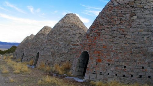 Four stone beehive-shaped kilns stand in a row against a blue sky, surrounded by dry grass and distant mountains.