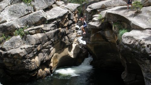 Two people near a rocky gorge with a flowing river, one jumping between rocks and the other standing on a ledge.