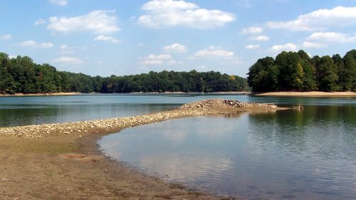 A serene lake surrounded by trees, with a rocky formation extending into the water under a blue sky with fluffy clouds.