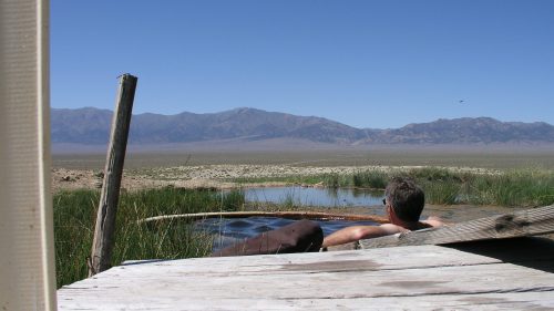 A person relaxes in a hot spring, surrounded by grass and mountains under a clear blue sky.