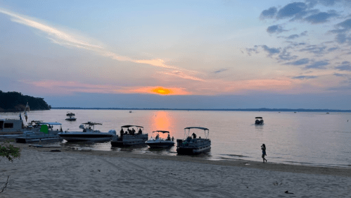 A serene beach at sunset with boats anchored and a person walking along the sandy shore.