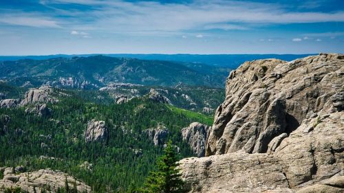 A panoramic view of rugged mountains and lush green forests under a blue sky with scattered clouds.