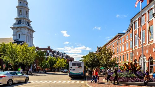 A sunny street scene featuring a church steeple, trees, and people walking near a brick building and an American flag.