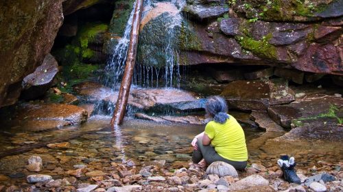 A person sits by a small waterfall surrounded by rocks and greenery, enjoying the serene natural setting.