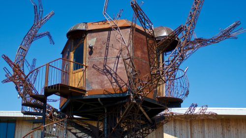 A large, rusted metal treehouse with a spiral staircase, set against a clear blue sky and modern building backdrop.
